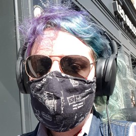 Photo of Enka Blanchard with multicolour hair, with their face hidden by sunglasses, a facemask and headphones.